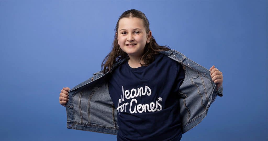 Young girl smiling in a Jeans for Genes T-shirt and denim jacket against blue background.