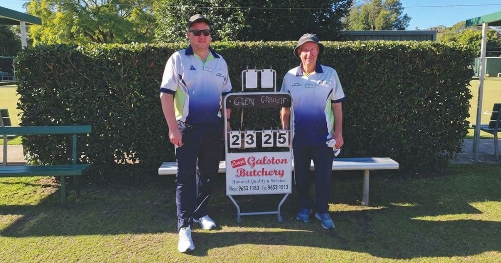 Two lawn bowl players standing outdoors with a scoreboard at Dural Bowling Club.