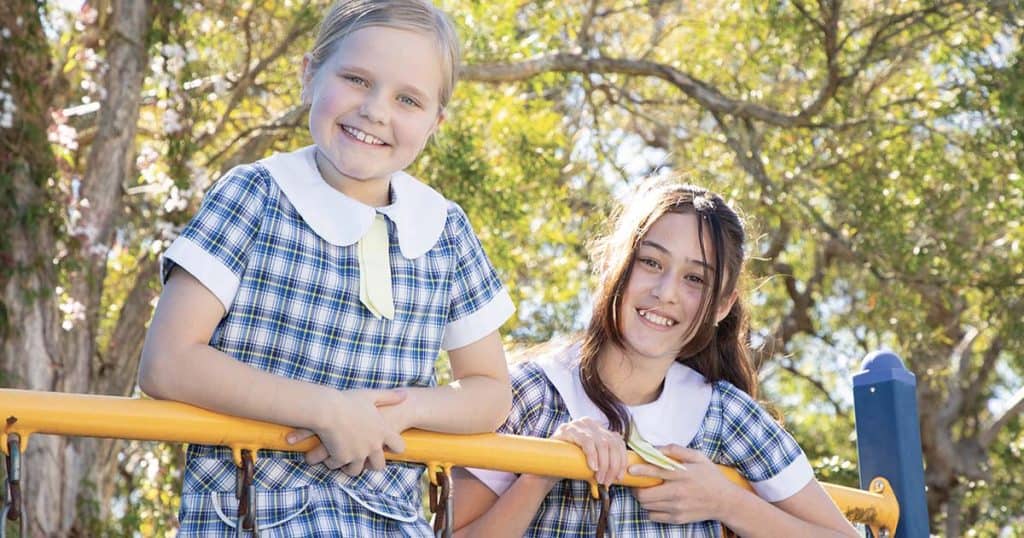 Two smiling schoolgirls in plaid uniforms on a sunny playground.